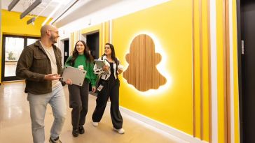 Snap team members smiling and talking while walking through office hallway featuring yellow wall with illuminated Snap logo