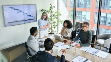 A businesswoman leads a group meeting with clients, discussing a presentation slide that is on a screen behind her. 