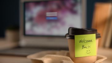 A yellow sticky note on a to-go coffee cup welcomes an employee back to the office.