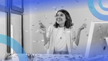 A business woman sitting at a computer desk, smiling with confetti raining down on her.