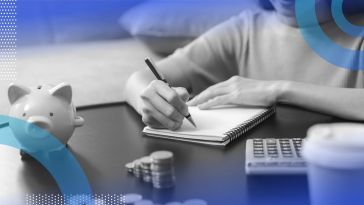 A close-up of a woman writing in a notebook with a piggy bank, stack of quarters, calculator and disposable coffee cup next to her.