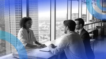 A woman being interviewed by two men at a table in a windowed conference room.