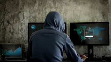 A photo of a person in a hoodie in front of computer monitors. 