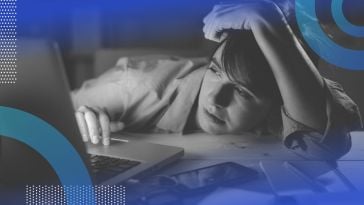 A young woman is slumped over her desk at work, staring wearily at her computer screen.