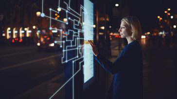 A woman stands in front of a futuristic transparent touch screen while selecting an option on a menu.