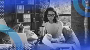 A young woman in glasses holding a coffee cup leafs through papers next to a laptop in an office full of books