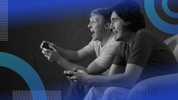 Two young men are holding gaming consoles and looking excitedly at a screen.