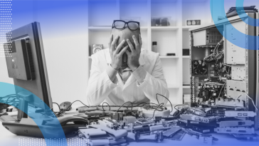 A frustrated man sitting at a desk littered with machine parts with his head in his hands and glasses on top of his head.