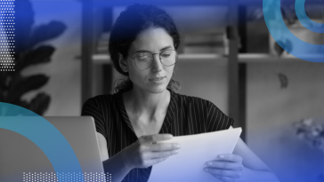 A woman at a desk with a laptop, considering a piece of paper.