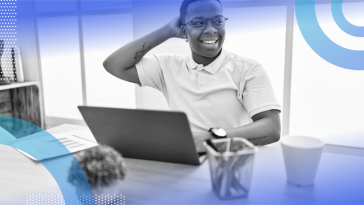 A young trans masc person smiling with one hand behind their head and one on their laptop keyboard.