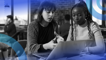 Two women are sitting at a desk discussing something on a computer screen.