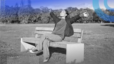 A smiling woman is sitting on a bench with her arms raised to the sky.