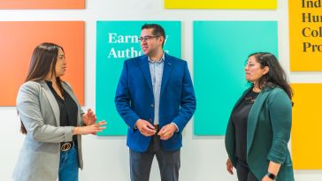 Three Udemy team members stand talking in front of a wall of colorful art.