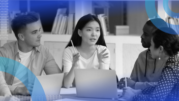 Four young professionals sitting at a desk with laptops having a discussion.