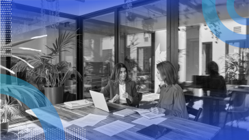 Two women talk at a table full of papers and computers