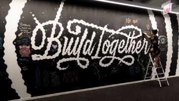 A woman stands on a step ladder to write with chalk on a large wall that reads “Build Together” in cursive and is decorated with chalk art.