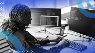 A woman from the waist up coding on a computer with two screens.