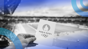 A hand is holding a card that says Paris 2024 in front of an athletic field.