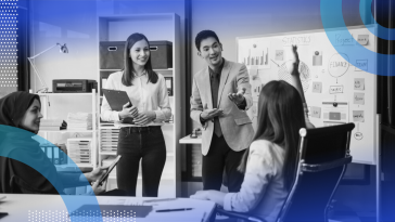 A young woman raising her hand in a meeting while team members smile at her and the leader calls on her.