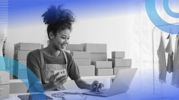 A smiling small business owner sitting at a desk on her laptop with her smart phone in the other hand and stacks of boxes behind her.