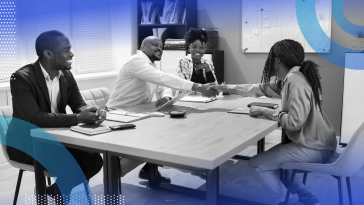 Two people are shaking hands at a conference table as two other people look on.