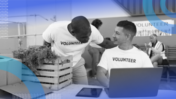 A person wearing a volunteer t-shirt is sitting at a computer and talking to another volunteer.