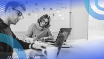 Two people are sitting at a desk working on a project. A whiteboard behind them is filled with sticky notes.