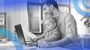  A man holding a baby is working from home with a laptop.