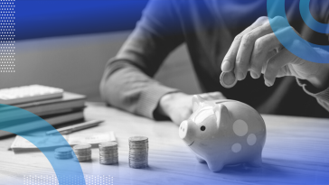 A person visible from the neck down and waist up sitting at a desk, adding a coin to a piggy bank next to inclining stacks of coins, dollar bills, a pen and a pile of notebooks.