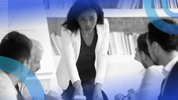 A woman professional standing to lead a meeting while her colleagues sit at the table in front of her.