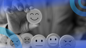 A close-up of a person from the shoulders down holding a wooden circle with a painted smiley face on it, with five other circles of wood sitting on the table with varying degrees of emotion painted on them.