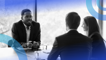 Three people are seated at a table. One is making a presentation to the other two.