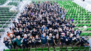  Large group of Iodine Software employees standing among seats in a stadium, cheering at camera. Photo taken from above. 