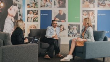 Three GoHealth employees sit in grey armchairs chatting with one another in front of a photo collage wall. 