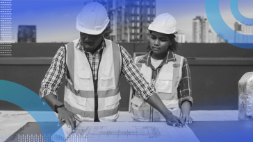 Two construction workers, one older and one younger, wearing safety vests and hard hats while looking over a blueprint together.