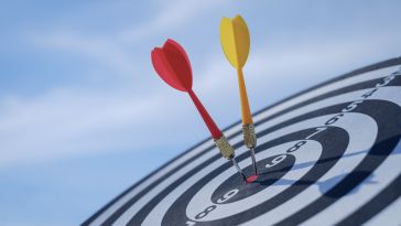 A close-up of a bullseye with two colored darts sticking out of the center circle against a blue sky background.