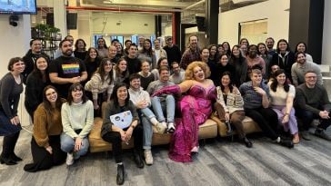 A group of Braze employees gathered for a group picture — some sitting on a couch and some standing — with a drag queen in a hot pink dress in the center. 