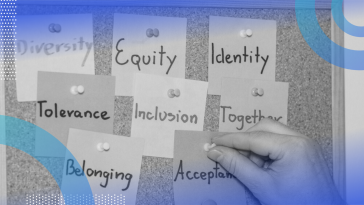 A close-up of a corkboard with sticky notes reading diversity, equity, identity, tolerance, inclusion, together, belonging and acceptance pinned to it. A person’s hand is actively pinning the acceptance note to the board.