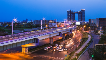 Cars zoom on Kolkata streets and highways at dusk with high-rises in the background.