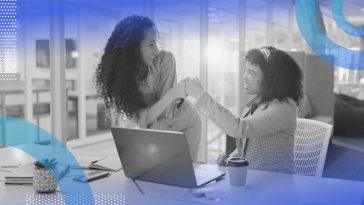 Two women colleagues, one sitting and one standing at a desk with an open laptop on it, fist bumping.