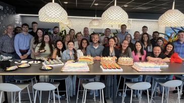 A group of McCain employees stand in a kitchen on one side of a long table covered in rainbow-colored cakes in celebration of Pride month. 