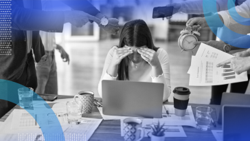 A woman hunched over her laptop shielding her face from several colleagues shoving clocks and papers at her.