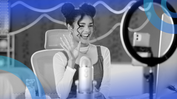 A smiling young woman wearing her hair in sports buns smiling and waving at her phone with a ring light behind it and microphone in front of her.