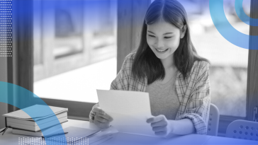 A woman smiling at a piece of paper while sitting next to a notebook, pen and book.