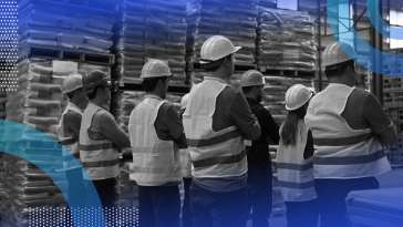 A group of workers in safety vests and hard hats stand in a warehouse