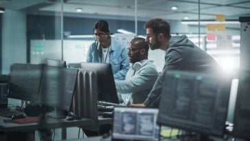 Three software engineers gathered around a computer monitor in an office, viewing work they’ve done on an AI project. 