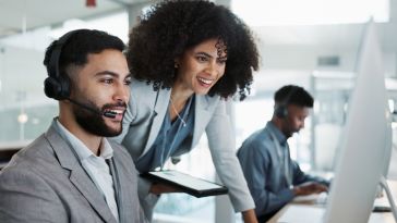 Colleagues looking at a monitor together, smiling. One is seated with a telephone headset, the other is standing next to him, leaning in. 