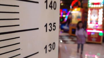 A photo of a defocused girl in the background with a pink shirt at an amusement park or game area, moving towards a wooden height board to measure minimum height requirements for ride.