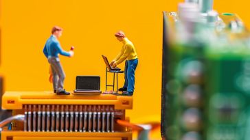 Two miniature figures working on a life-size computer circuit in front of a yellow background.