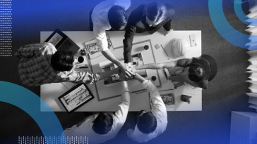 An aerial view of a team of six colleagues sitting at a table working on a project with their hands piled on top of one another’s in the center.